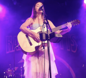 A woman in a white dress singing and playing an acoustic guitar.