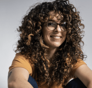 A woman with curly hair sitting on a grey background.