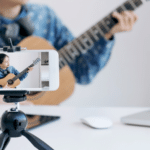 A woman is playing an acoustic guitar on a tripod in front of a laptop.