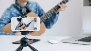 A woman is playing an acoustic guitar on a tripod in front of a laptop.