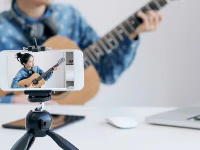 A woman is playing an acoustic guitar on a tripod in front of a laptop.