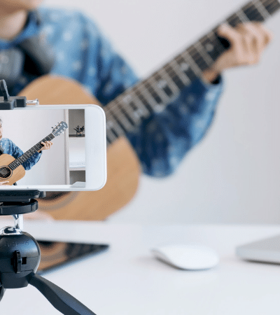 A woman is playing an acoustic guitar on a tripod in front of a laptop.