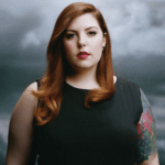 A woman with tattoos standing in front of a cloudy sky.