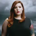 A woman with tattoos standing in front of a cloudy sky.