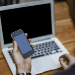 A man holding a cell phone in front of a laptop.