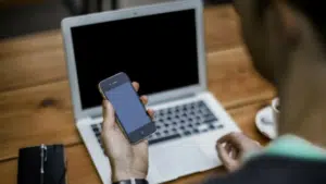 A man holding a cell phone in front of a laptop.