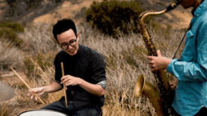 Two men playing saxophone and drums in a field.