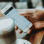 A hand holding a smartphone next to a cup of coffee.