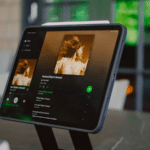 A tablet on a stand displays a Spotify music streaming app, showcasing album artwork featuring a person in motion.