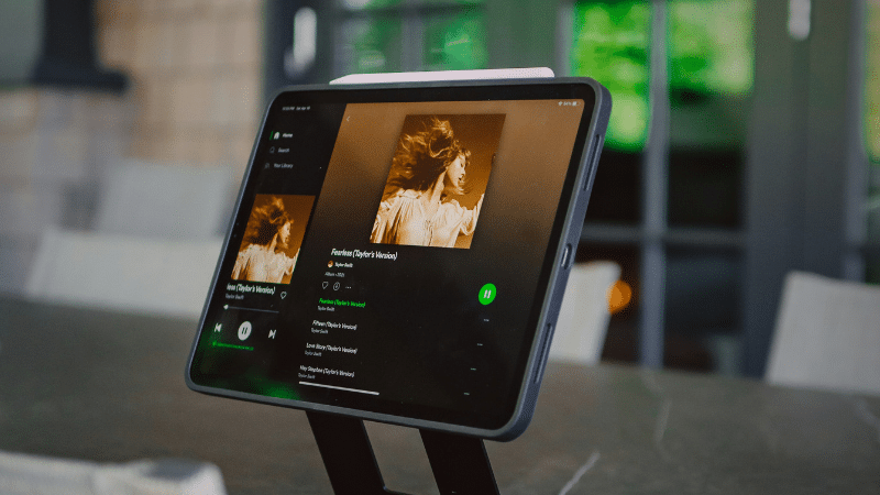 A tablet on a stand displays a Spotify music streaming app, showcasing album artwork featuring a person in motion.