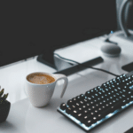 A desk with a computer, keyboard and cup of coffee.