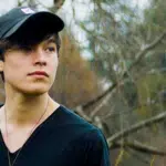 A young man in a black hat standing in a wooded area.