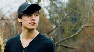 A young man in a black hat standing in a wooded area.