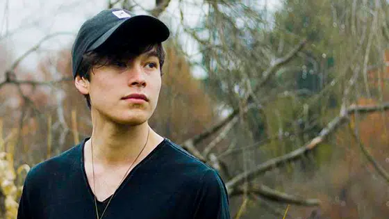 A young man in a black hat standing in a wooded area.