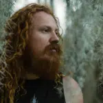 A man with long red hair standing in front of moss.