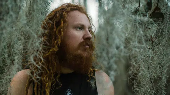 A man with long red hair standing in front of moss.