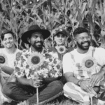 A black and white photo of a group of men sitting in front of a corn field.