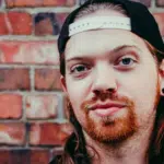 A man with long hair and a hat in front of a brick wall.