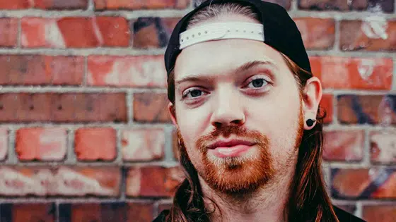 A man with long hair and a hat in front of a brick wall.