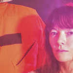 A woman in an orange jacket standing next to a mannequin.