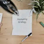 A notepad with the word marketing strategy on it sits on a wooden table.