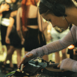 A DJ with headphones operates a mixer at a lively market event, with blurred people in the background.