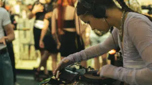 A DJ with headphones operates a mixer at a lively market event, with blurred people in the background.