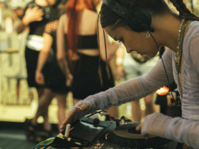 A DJ with headphones operates a mixer at a lively market event, with blurred people in the background.