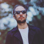 A man in a black jacket and sunglasses is standing in front of trees.