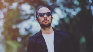 A man in a black jacket and sunglasses is standing in front of trees.