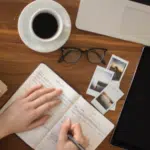 A woman's hand writing on a notebook with a cup of coffee and a laptop.