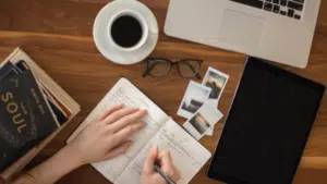 A woman's hand writing on a notebook with a cup of coffee and a laptop.