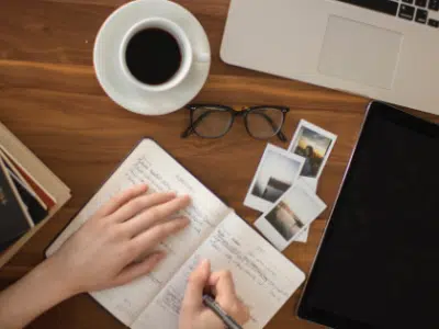 A woman's hand writing on a notebook with a cup of coffee and a laptop.