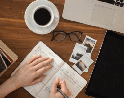 A woman's hand writing on a notebook with a cup of coffee and a laptop.