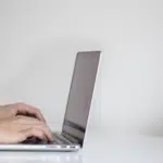 A man typing on a laptop in front of a potted plant.