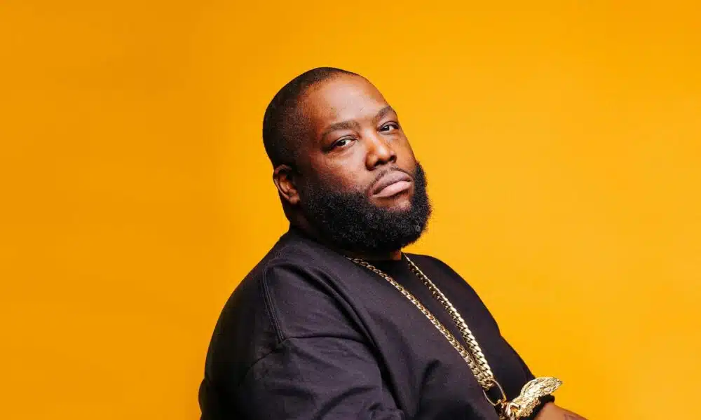 A black man with a beard sitting on a yellow background.