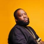 A black man with a beard sitting on a yellow background.