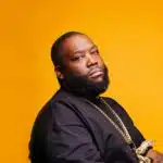 A black man with a beard sitting on a yellow background.