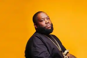 A black man with a beard sitting on a yellow background.