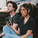 A young couple sitting next to a car listening to music.