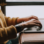 A woman typing on a laptop in front of a window.