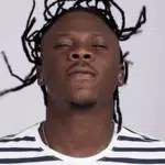 A man with dreadlocks is standing in front of a white background.