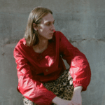 A woman in a red jacket and leopard print pants leaning against a wall.