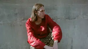 A woman in a red jacket and leopard print pants leaning against a wall.