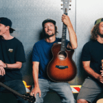 Three men are posing with guitars in front of a metal wall.