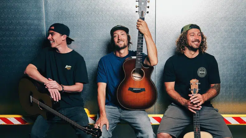 Three men are posing with guitars in front of a metal wall.