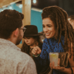 Several people socialize at an outdoor event with lively Latin music playing in the background. A woman with long dreadlocks holds a drink while talking to a man.