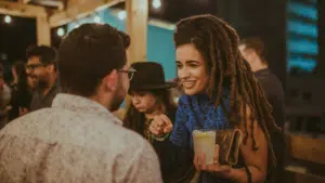 Several people socialize at an outdoor event with lively Latin music playing in the background. A woman with long dreadlocks holds a drink while talking to a man.