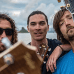 Three men are holding guitars in front of a body of water.