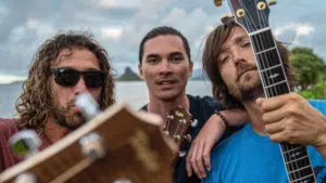 Three men are holding guitars in front of a body of water.